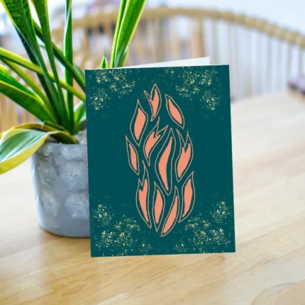 Glittering celebration greeting card featuring a central abstract flame or leaf design in peach on a deep teal background, accented with gold sparkle effect. The card is displayed back vase of plant against a vibrant blue background.