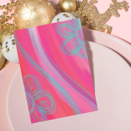 Close-up of a vertical hot pink and teal abstract greeting card featuring sketchy floral outlines, resting on a light pink plate and surrounded by festive gold and white Christmas ornaments and gold beaded garlands.