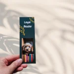 Cute magnetic dog bookmark featuring a brown and white Cocker Spaniel puppy illustration peeking over a stack of colorful books on a dark teal background