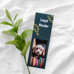 Cute magnetic dog bookmark featuring a brown and white Cocker Spaniel puppy illustration peeking over a stack of colorful books on a dark teal background, resting on a white sheet next to a green plant.
