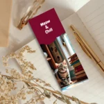 Cute tabby cat kitten sitting next to stacked books on a wooden floor in front of a sunny window on a maroon bookmark with the text Meow & Chill. Perfect reading accessory