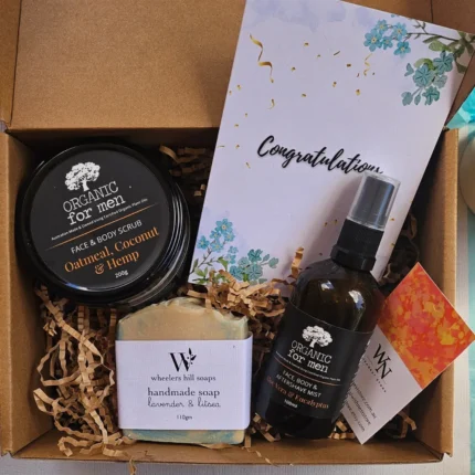 Organic men's grooming gift box featuring oatmeal face scrub, handmade lavender soap, eucalyptus aftershave mist, and a congratulations card on a kraft paper background.