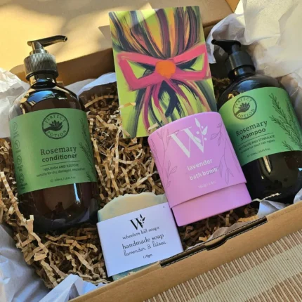 An open eco-friendly kraft gift box filled with Perfect Potion Rosemary Shampoo and Conditioner, Wheelers Hill Lavender soap, and a purple bath bomb on a bed of recycled paper shred.