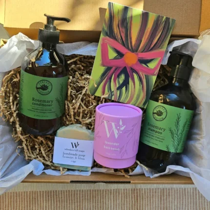 An open eco-friendly kraft gift box filled with Perfect Potion Rosemary Shampoo and Conditioner, Wheelers Hill Lavender soap, and a purple bath bomb on a bed of recycled paper shred.