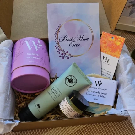 An open WishNest Store Mother's Day gift box on a wooden surface, featuring Perfect Potion Calendula hand cream, Rose Petals lip balm, a lavender bath bomb, handmade lavender and litsea soap, and a "Best Mom Ever" floral card.