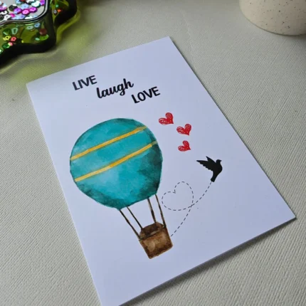 A white greeting card featuring a teal watercolor hot air balloon and the text Live Laugh Love.