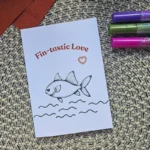A vertical greeting card with a hand-drawn fish illustration and the text "Fin-tastic Love" on a woven mat.