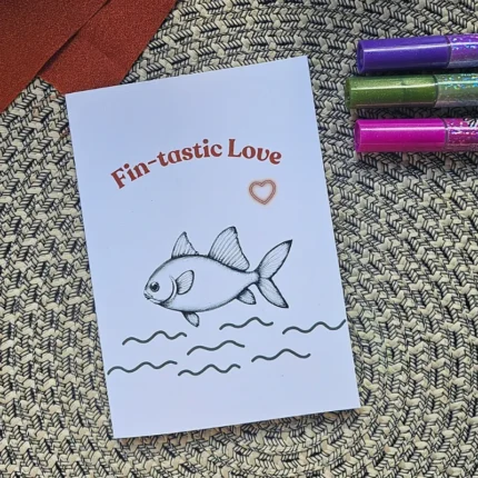 A vertical greeting card with a hand-drawn fish illustration and the text "Fin-tastic Love" on a woven mat.