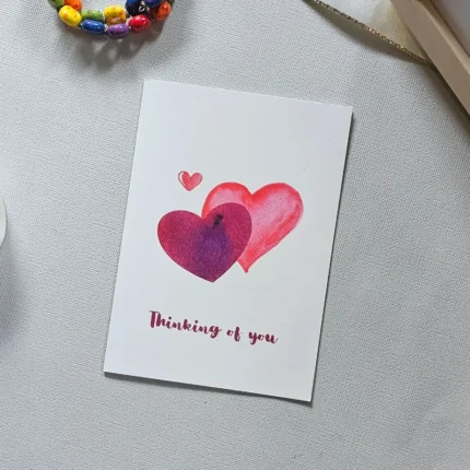 A vertical "Thinking of You" greeting card with two watercolor hearts on a grey textured surface.
