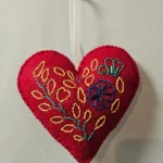 A close-up shot of a vibrant red, hand-embroidered felt heart keepsake ornament featuring intricate yellow, blue, and green floral stitching with an attached white satin ribbon against a textured off-white background.