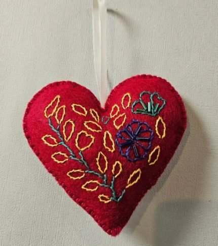 A close-up shot of a vibrant red, hand-embroidered felt heart keepsake ornament featuring intricate yellow, blue, and green floral stitching with an attached white satin ribbon against a textured off-white background.