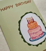 Close-up of the illustrated pink and orange two-tier birthday cake on the sage green birthday card