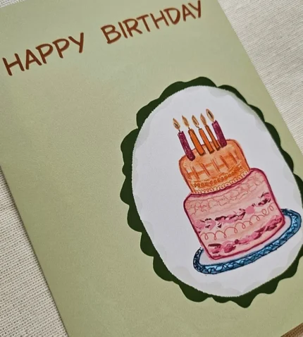 Close-up of the illustrated pink and orange two-tier birthday cake on the sage green birthday card
