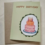 Hand-drawn birthday cake card with five candles on a sage green Happy Birthday card, kraft envelope behind